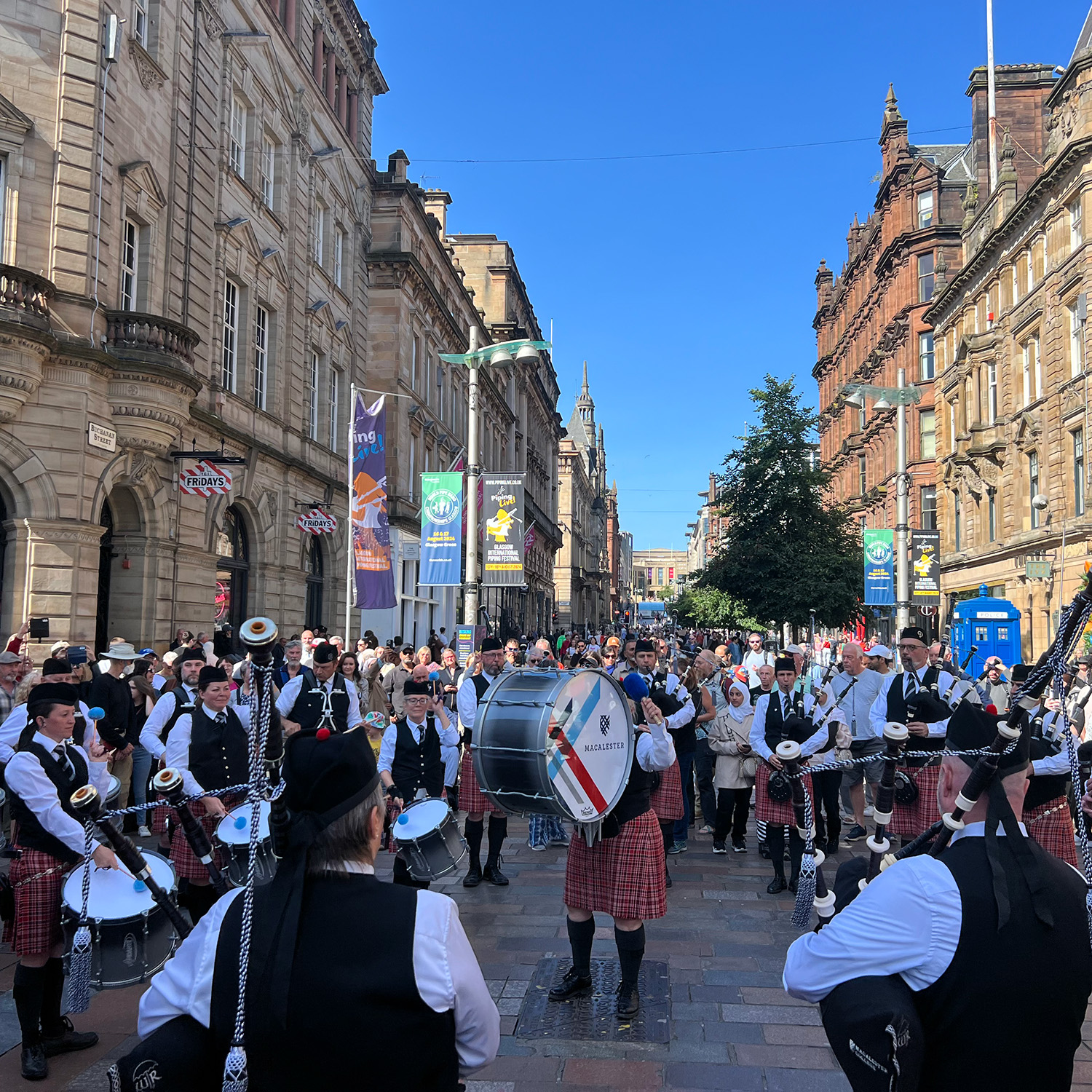 The Macalester Pipe Band performs on a street in Scotland. The group is standing in a circle with a drummer in the middle. Old white brick buildings are on both sides of the street.