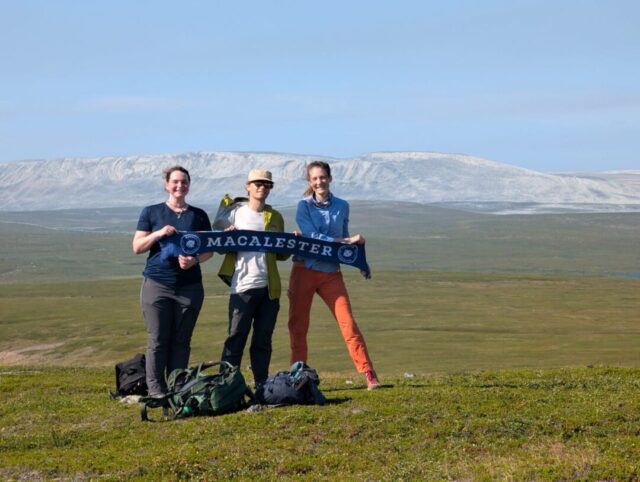 Lukas Lock-Scamp ’25, Mary Ossar ’24, and biology professor Mary Heskel pose with a Macalester banner while standing on green grass in Arctic Norway.