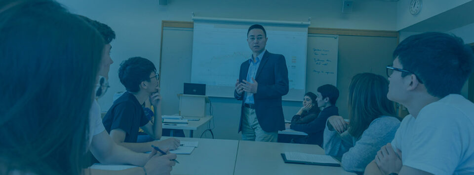 A professor stands in front of a screen teaching a class as seven students sitting at desks watch and write in notebooks