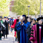 A row of professors standing on campus in academic regalia; one claps and smiles