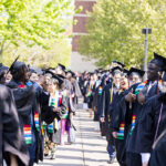 Two rows of students in graduation gowns and caps walk along the sidewalk parallel to each other; two students give each other thumbs up
