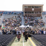 Two bagpipers lead two rows of students down an aisle between chairs on a football field