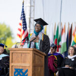 Commencement speaker Anna Sene stands at a podium, smiling with her hand over her heart
