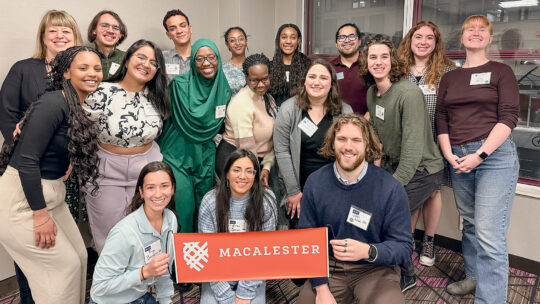Seventeen students and Macalester staff members pose for a picture holding a Macalester banner
