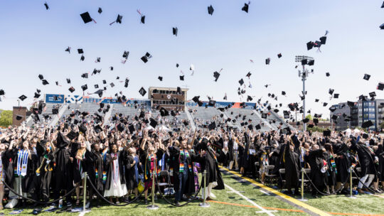 A large group of students in black graduation gowns stands on the field of Macalester Stadium, throwing their caps into the air and smiling