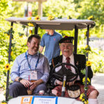 A man wearing a traditional kilt drives a golf cart carrying two Reunion attendees