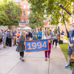 A parade of alumni walk down a path, following two alumni carrying a banner reading 1994