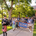 A parade of alumni walk down a path, following two alumni carrying a banner reading 2014 and a bagpiper