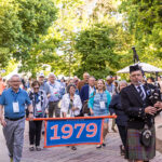 A parade of alumni walk down a path, following two alumni carrying a banner reading 1979 and a bagpiper