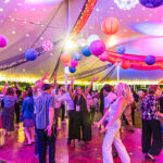 A group of attendees dance on a dance floor under an outdoor tent decorated with blue and orange streamers