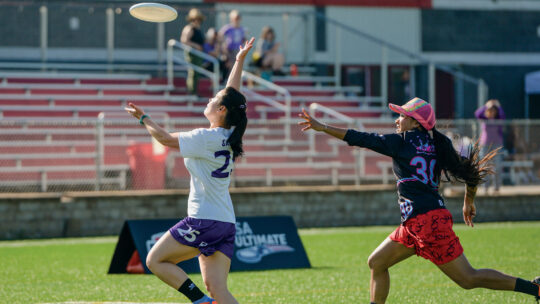 A Macalester Frisbee player and a player on the opposing team reach into the air to grab a Frisbee