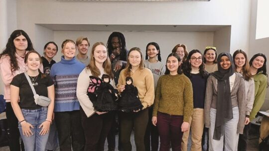 Members of English professor Andrea Kaston Tange's nineteenth-century British literature class posing with two dolls that they sewed dresses for.