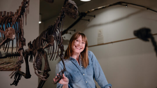 Kristi Curry Rogers stands in front of a displayed dinosaur skeleton