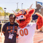 An alum smiles with an orange Highland cow mascot (Coo) as somebody else takes their picture with a smartphone