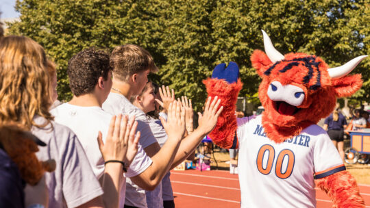 An orange Highland cow mascot (Coo) high-fives a row of students on a track field