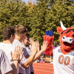 An orange Highland cow mascot (Coo) high-fives a row of students on a track field