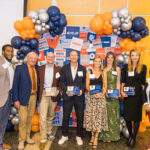 2024 Hall of Fame inductees hold plaques and stand against a balloon arch with President Suzanne Rivera, Athletics Director Donnie Brooks, and M Club President Steve Cox