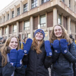 Three Mac students stand outside, holding up their hands to display new Mac branded athletics mittens.