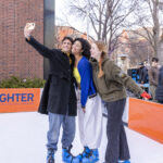 One Mac student holds up a phone to take a selfie with two other students. They're all wearing ice skates and standing on the ice rink at the Brighter campaign launch event.