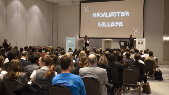 Attendees and participants of the National Ethics Bowl Championship sit in chairs in a large conference room. The screen on the wall reads 