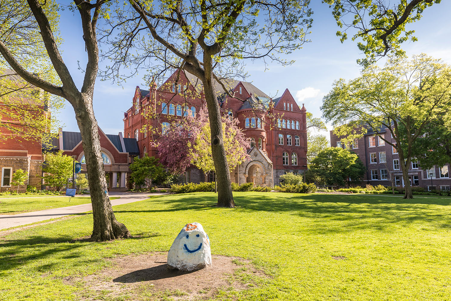 The rock on Macalester's Great Lawn is painted with a white smiley face and orange heart. Old Main is visible in the background.