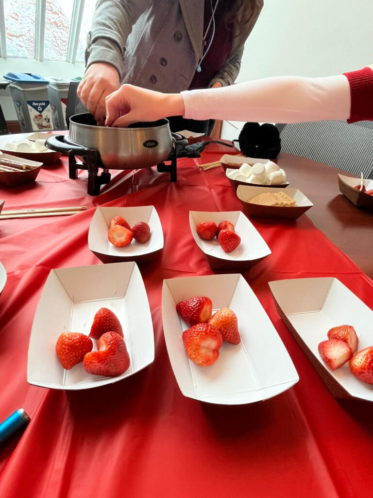 Five paper food boat trays with strawberries in them on a red tablecloth at the Valentine's Day coffee house. Behind them on the table, a heater with chocolate fondue inside and two hands reaching out to dip food into it.