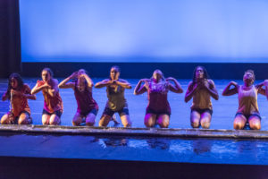 Several dancers kneel on stage in a line