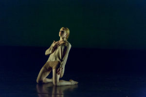 Dancer kneels on stage, holding a hand to their chest