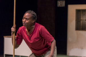 Actor wearing red crouches and holds the handle of a mop