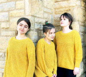 Three performers wearing yellow sweaters stand in front of a light brick wall