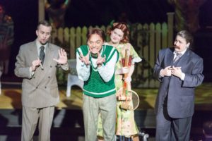 Actor in a green vest makes a grotesque face as three others stand around watching