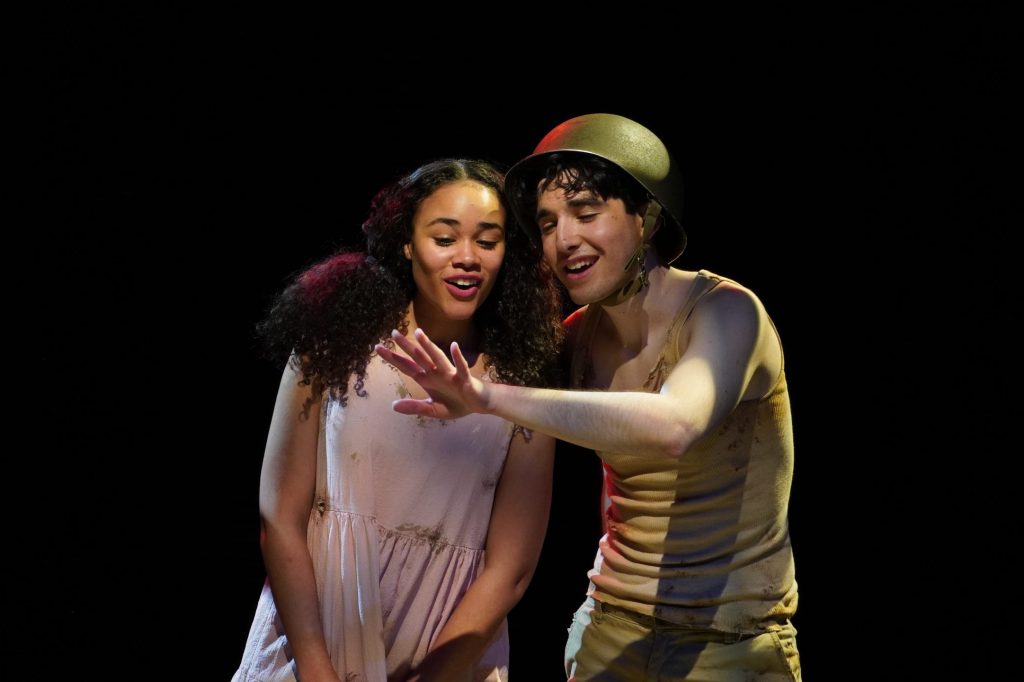 two actors stand on a dark stage, admiring a ring on one of their hands.